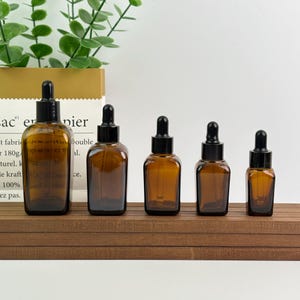 May include: Five amber glass dropper bottles of varying sizes are arranged on a wooden shelf. Each bottle has a black dropper top. The bottles are in front of a white background and some green leaves.
