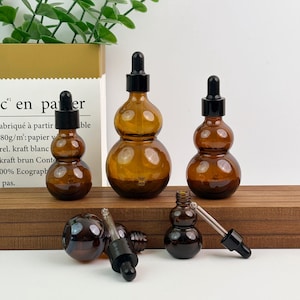 May include: Amber glass dropper bottles in various sizes, with black dropper tops. The bottles have a unique, gourd-like shape. Several bottles are arranged on a wooden surface, with a paper bag in the background.