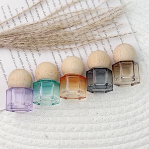 Könnte beinhalten: Eine Reihe von fünf farbigen Glas-Diffusorflaschen für ätherische Öle mit Holzdeckeln. Die sechseckigen Flaschen sind lila, türkis, orange, grau und klar. Trockenes Gras und ein Papier mit Text im Hintergrund.