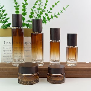 May include: A collection of four amber glass spray bottles and two amber glass jars, all featuring dark brown wooden caps. The bottles vary in height, while the jars are short and wide with faceted bases, creating a uniform set of containers.