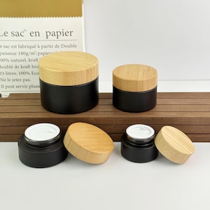 May include: Black cosmetic jars with bamboo lids. The jars are cylindrical with a matte black finish. The lids are made of light-colored bamboo. Some jars have their lids off, revealing a white interior.