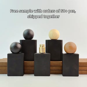 May include: A display of black rectangular perfume bottles with wooden sphere toppers. One bottle has a gold-colored atomizer. The bottles are arranged on a wooden platform. The text "Free sample with orders of 50+ pcs, shipped together" is visible.