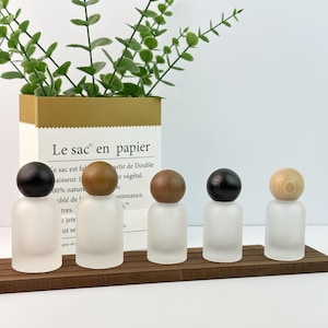May include: Five frosted glass bottles with wooden ball-shaped stoppers in varying shades of brown and black. The bottles are arranged on a dark brown wooden base. A paper bag with the text "Le sac en papier" is in the background.