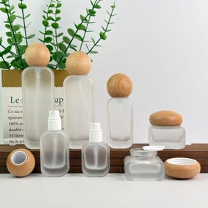 May include: A collection of frosted glass bottles and jars with wooden lids and spray nozzles. The bottles vary in size and shape, with a rectangular and rounded design. The wooden lids are light brown. The image is set against a white background with green foliage.