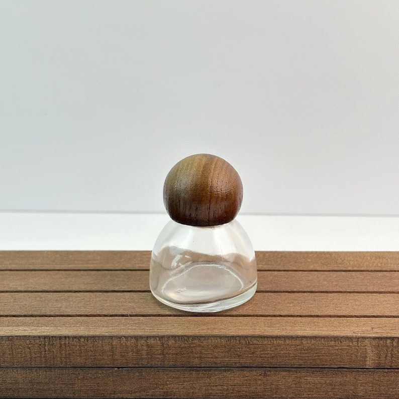 May include: A clear glass bottle with a rounded wooden stopper. The bottle is empty and sits on a brown wooden surface. The stopper is a dark brown color and is spherical in shape. The background is white.