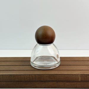 May include: A clear glass bottle with a rounded base and a dark brown wooden ball stopper. The bottle sits on a brown wooden surface against a white background. The design is simple and elegant.
