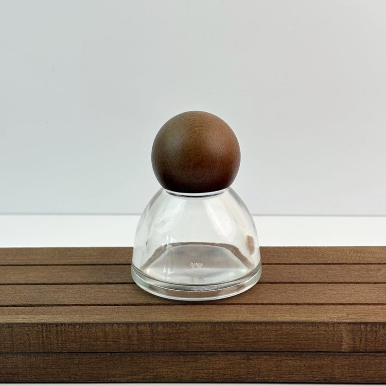 May include: A clear glass bottle with a rounded base and a dark brown wooden ball stopper. The bottle sits on a brown wooden surface against a white background. The design is simple and elegant.