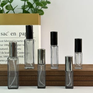 Op de afbeelding: Een verzameling rechthoekige glazen parfumflesjes met zwarte en zilveren doppen. De flesjes staan op een houten oppervlak, met een papieren zak op de achtergrond. De flesjes zijn leeg en klaar om gevuld te worden.