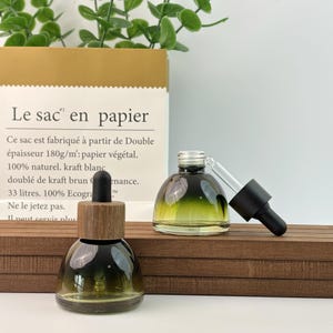 May include: Two glass dropper bottles with a gradient green and clear design. One bottle has a wooden cap, the other has a black cap. The bottles sit on a wooden surface, with a paper bag in the background.