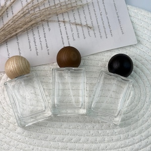 May include: Three clear glass perfume bottles with wooden and black spherical caps are arranged on a white woven surface. The bottles are rectangular with a spray nozzle. A sprig of dried grass and a piece of paper with text are in the background.
