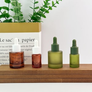 May include: Two amber glass dropper bottles with white caps and two matte olive green glass dropper bottles with matching green caps. The bottles are arranged in a row on a wooden surface in front of a paper bag with French text printed on it.