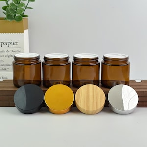 Puede incluir: Se muestran cuatro frascos de vidrio ámbar con tapas blancas, cada uno con un estilo de tapa diferente. Las tapas incluyen negro, dorado, veta de madera y plateado. Los frascos son cilíndricos y se asientan sobre una superficie de madera.