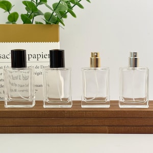 Peut inclure: Quatre flacons de parfum en verre transparent avec différents vaporisateurs. Deux flacons ont des bouchons noirs, un a un vaporisateur doré et le dernier a un vaporisateur argenté. Les flacons sont rectangulaires et reposent sur une surface en bois.