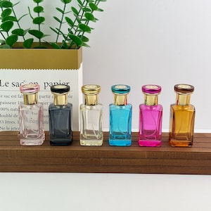 May include: Six rectangular glass perfume bottles with gold-toned caps, arranged in a row. The bottles are colored light pink, black, clear, bright blue, magenta, and amber. Each bottle features a clear spray nozzle and a gold-toned collar.