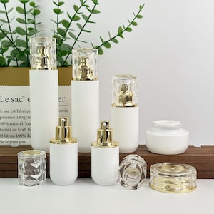 Peut inclure: Ensemble de flacons et pots cosmétiques blancs avec des détails dorés. Les flacons ont des bouchons facettés transparents et des vaporisateurs. Les pots et flacons sont disposés sur une surface en bois, avec du feuillage vert en arrière-plan.