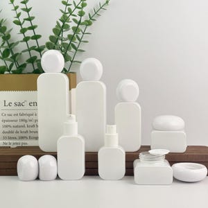 May include: A collection of white cosmetic bottles and jars of various shapes and sizes. The containers include bottles with rounded tops, spray bottles, and jars with lids. The items are arranged on a wooden surface.