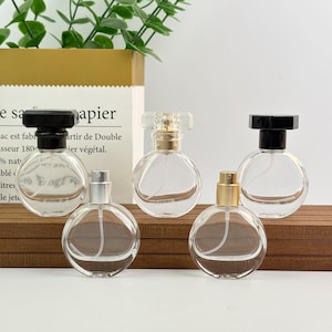 May include: Five clear, round perfume bottles with various cap designs. The caps are black, silver, gold, and clear. The bottles are displayed on a wooden surface with a paper bag and greenery in the background.