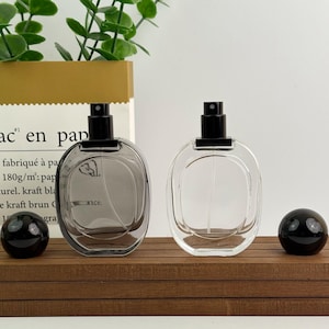 May include: Two oval-shaped perfume bottles with black spray nozzles. One bottle is clear, the other is a smoky gray. Two black spherical stoppers sit on a wooden surface. The background includes a paper bag with French text and green foliage.