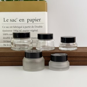 May include: A collection of five empty cosmetic jars with black screw-on lids. Three jars are made of clear glass, while two jars are made of frosted glass. The jars are arranged in two rows on a wooden surface in front of a paper bag.