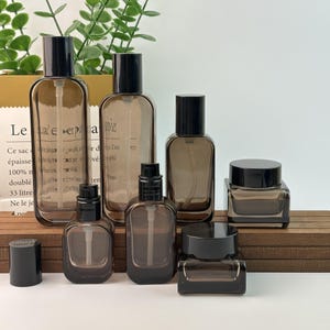 May include: A selection of brown glass bottles and jars with black lids. The set includes different sizes of spray bottles and jars, displayed on a wooden surface. The bottles have a rectangular shape with rounded corners.