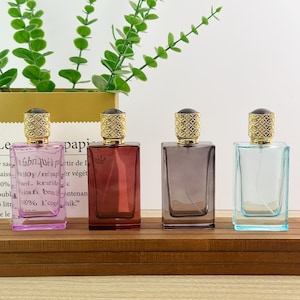 May include: Four rectangular glass perfume bottles with gold-toned textured caps arranged in a row. The bottles are colored light purple, deep red, dark gray, and light blue. Each bottle features a clear spray nozzle and a transparent glass body.