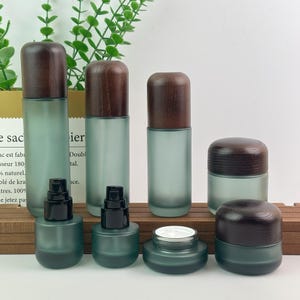 Peut inclure: Un ensemble de contenants cosmétiques en verre dépoli vert avec des couvercles en bois brun foncé. Le set comprend trois flacons pompe hauts, deux flacons pompe courts et deux pots larges avec des bouchons en bois assortis sur un socle.