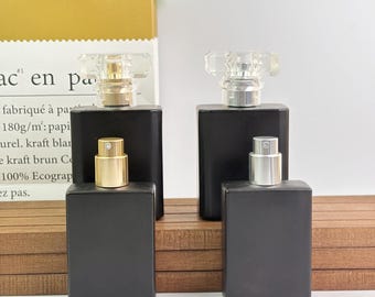 May include: Four black rectangular perfume bottles with gold and silver sprayers and clear, faceted tops. The bottles are arranged on a wooden surface, with a paper bag in the background.
