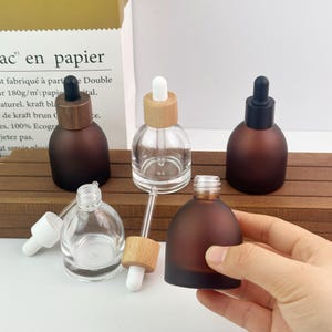 May include: A collection of round glass dropper bottles with various finishes, including frosted brown and clear glass. The bottles feature wooden or black screw-top caps with integrated glass droppers. Some bottles are assembled, while others are shown with the dropper removed, displaying the threaded neck.