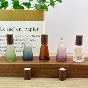 May include: Five small glass roller bottles with dark wood caps. Three bottles have a frosted finish in light green, dark blue, and purple. One bottle is clear amber glass, and one is clear glass. Two extra wood caps sit on the wooden surface.