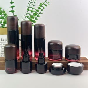 Könnte beinhalten: Eine Sammlung von Kosmetikbehältern mit dunkelroten Glasflaschen und Tiegeln mit dunkelbraunen Holzmaserungsdeckeln. Das Set umfasst hohe Pumpflaschen, kürzere Pumpflaschen und breite Tiegel, teilweise ohne Deckel mit weißen Einsätzen.