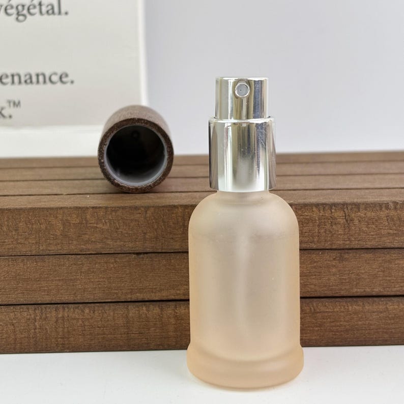 May include: A frosted glass perfume bottle with a silver spray top and a separate brown cap. The bottle has a rounded shape and a soft, peach-coloured hue. The cap is made of a dark, textured material. The bottle is designed for fragrance or cosmetic products.