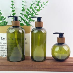 Puede incluir: Tres dispensadores de jabón de color verde oliva translúcido con mecanismos de bomba negros y collares de madera. Dos dispensadores son altos y cilíndricos, mientras que el tercero es una botella esférica más pequeña con un tubo interno.