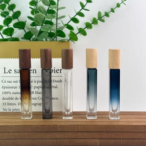 May include: Five rectangular glass perfume bottles with wooden caps arranged in a row. The bottles feature gradient colors ranging from dark brown to clear and blue. Each bottle has a cylindrical wooden lid and a slender, tall, clear glass body.