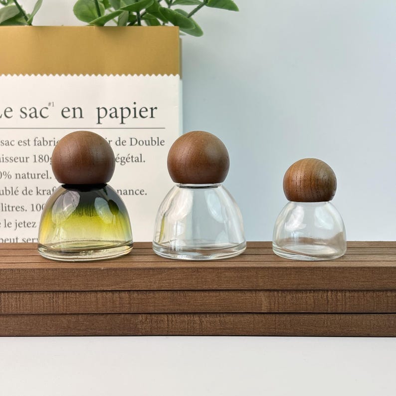 May include: Three glass bottles with wooden spherical stoppers. The bottles are clear, with one having a gradient of yellow and green. They sit on a brown wooden surface, with a paper bag in the background.