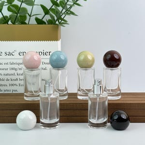 May include: Clear glass perfume bottles with silver sprayers and colorful spherical stoppers. The stoppers are pink, blue, yellow, and brown. There are also white and black stoppers. The bottles are displayed on a wooden surface.