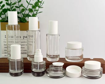 May include: A collection of clear glass cosmetic containers with white lids. The assortment includes various sizes of bottles, jars, and a spray bottle. The containers are arranged on a wooden surface, with a paper bag and greenery in the background.