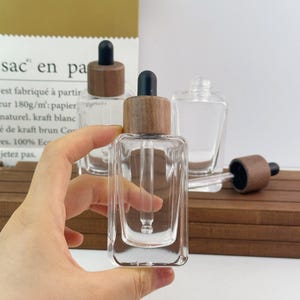 May include: A set of clear glass dropper bottles with square bases and wooden collars. Each bottle features a black rubber bulb and a glass pipette. The bottles are transparent, showcasing a minimalist design with a natural wood finish on the cap.
