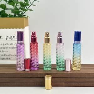 May include: Five colorful glass perfume bottles with matching caps. The bottles are in shades of purple, red, yellow, green, and blue. Each bottle has a ribbed design and a spray nozzle. A gold-colored bottle cap sits in front.