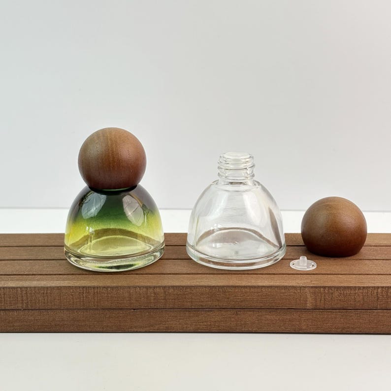 May include: Clear glass perfume bottle with a wooden cap, next to a similar bottle without a cap. The bottle on the left has a gradient of green and yellow. The bottles are on a wooden surface.