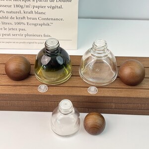 May include: Three glass perfume bottles with wooden spherical stoppers. One bottle has a gradient green tint, another is clear, and the third is also clear. Small plastic inserts are also present. The bottles are displayed on a wooden surface.