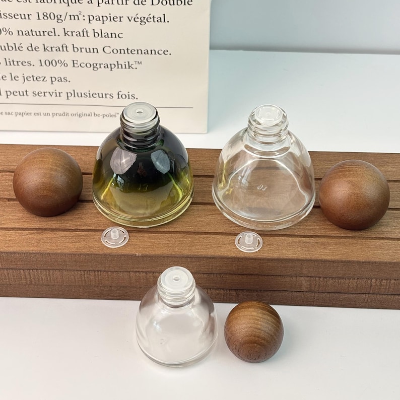 May include: Three glass perfume bottles with wooden spherical stoppers. One bottle has a gradient green tint, another is clear, and the third is also clear. Small plastic inserts are also present. The bottles are displayed on a wooden surface.