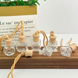 Può includere: Bottiglie di aromaterapia in vetro trasparente con tappi in legno e cordini beige disposte su una superficie di legno. Le bottiglie hanno forme diverse, tra cui rotonde, quadrate e un design a rosa. Sullo sfondo, un sacchetto di carta con testo francese.