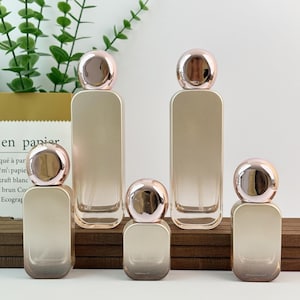 Könnte beinhalten: Fünf Parfümflaschen mit goldfarbenem Farbverlauf und metallisch roségoldenen Kappen. Die Flaschen stehen auf einer Holzoberfläche. Im Hintergrund ist Grün zu sehen. Jede Flasche hat eine rechteckige Form.