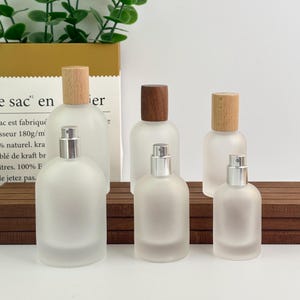 May include: A collection of frosted glass perfume bottles with silver sprayers and wooden caps. The bottles vary in size and are arranged on a wooden surface. The background includes a paper bag with French text and green foliage.