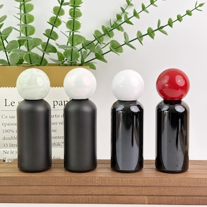 May include: Four decorative bottles with spherical stoppers. The bottles have black bodies and white or red stoppers. The bottles are arranged on a wooden surface, with greenery in the background.