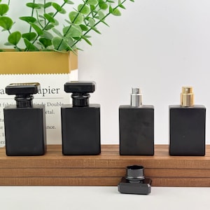 May include: Four black rectangular perfume bottles with different cap styles are displayed on a wooden surface. One bottle has a black square cap, another has a silver sprayer, and a third has a gold sprayer. A detached black cap sits in front.