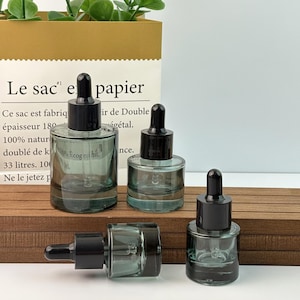 May include: Four small, clear glass dropper bottles with black caps are arranged on a wooden surface. The bottles are filled with a light green liquid. The background includes a paper bag with French text.