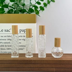 May include: Four clear glass roller bottles with light brown wooden caps. Three bottles are tall with rectangular bases and textured geometric patterns on the glass, while the fourth bottle is short and spherical with a smooth glass surface.