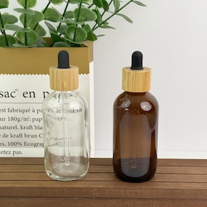 May include: Two glass dropper bottles with bamboo lids. One bottle is clear, and the other is amber-colored. Both bottles have a black dropper and are sitting on a wooden surface. The background includes green foliage and a paper bag.