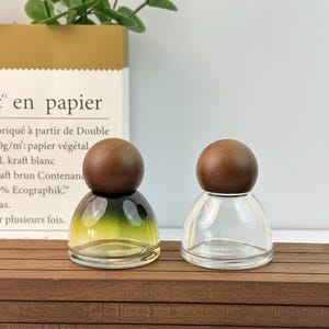 May include: Two small glass bottles with wooden spherical stoppers. One bottle has a gradient green and yellow tint, while the other is clear. The bottles sit on a brown wooden surface.